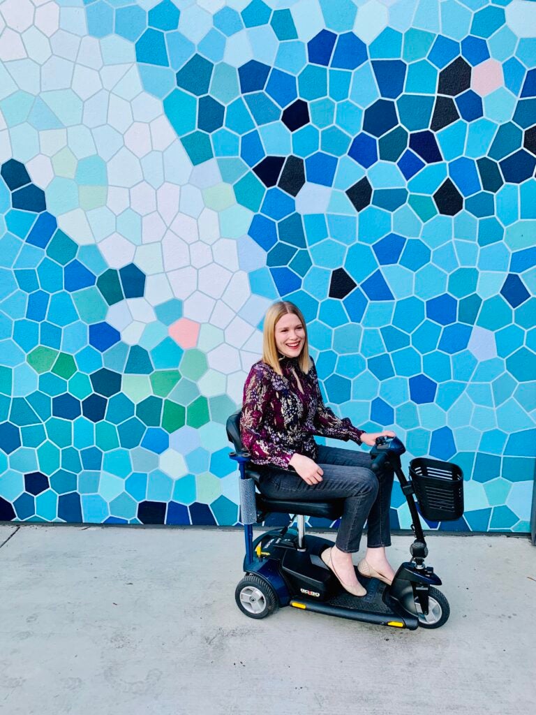 Amy, a white woman with straight, shoulder-length blonde hair, wears a burgundy blouse with gray jeans and sits in her mobility scooter in front of a wall with blue and white geometric shapes. 