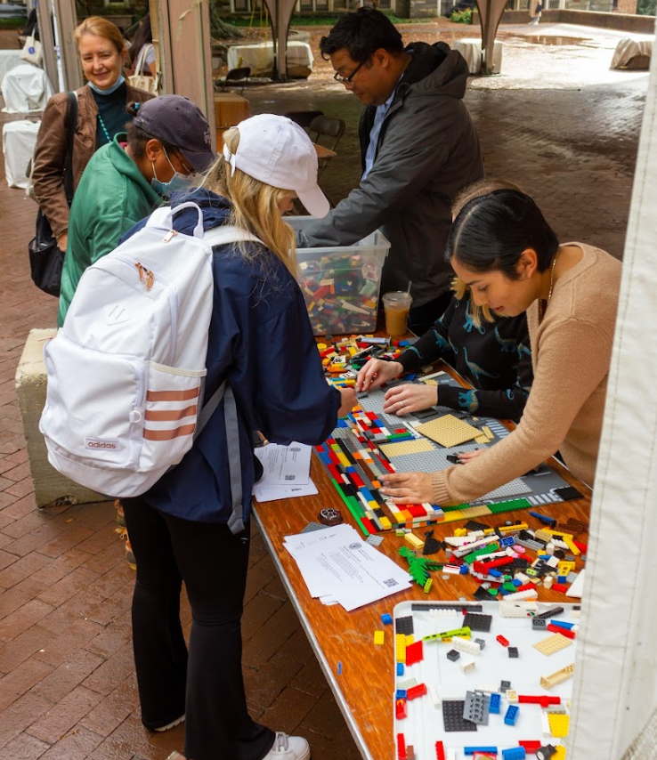 In Red Square, we have 6 people: 4 faculty from across campus and 2 student/student workers, working on the lego ramp. There are legos spread across the table along with someone going through the bucket of legos. The foundation of the platform for the ramp is done and the staircases are developing.