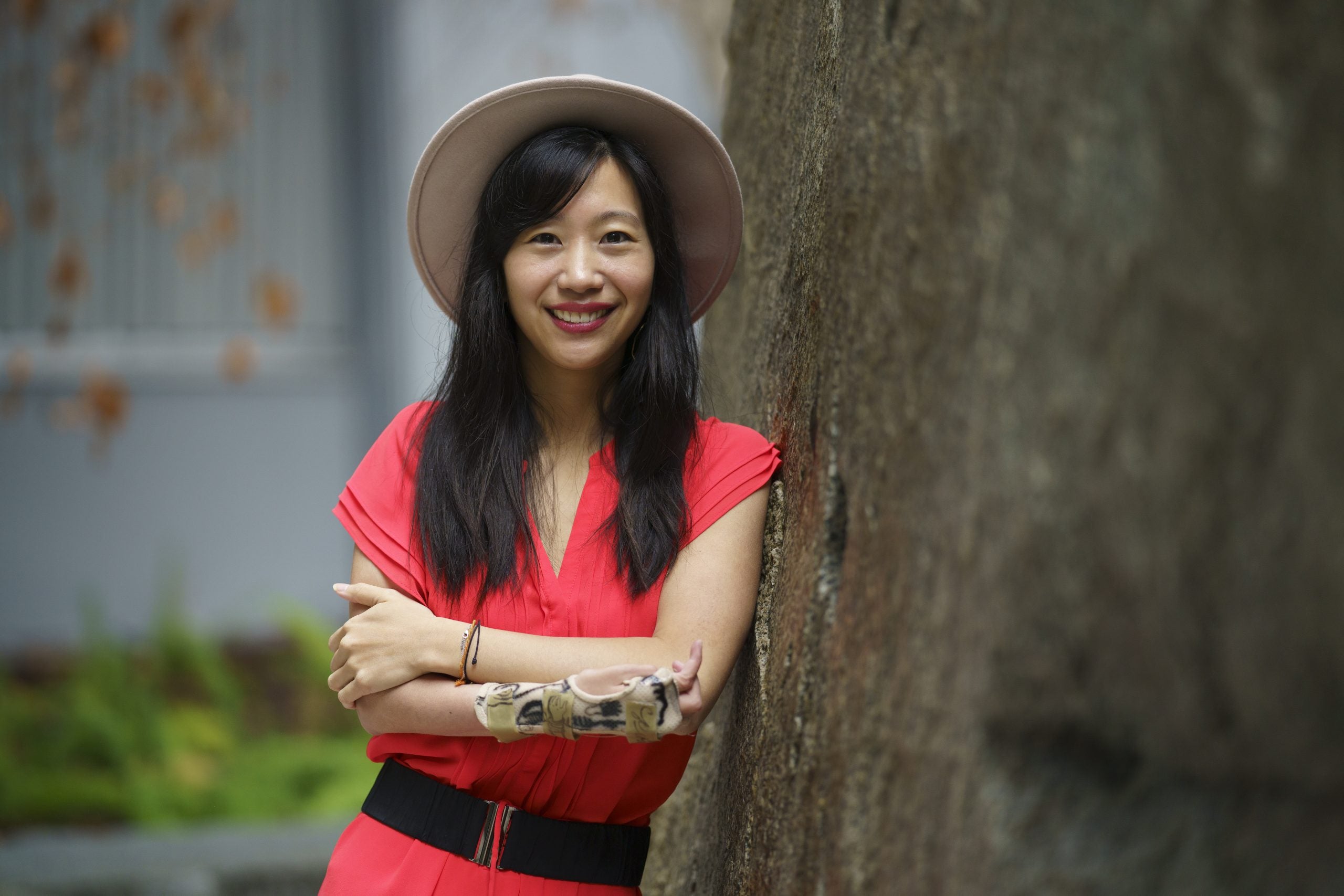 A picture of Tiffany Yu earring a red dress, black belt, and beige hat, leaning on a tree.