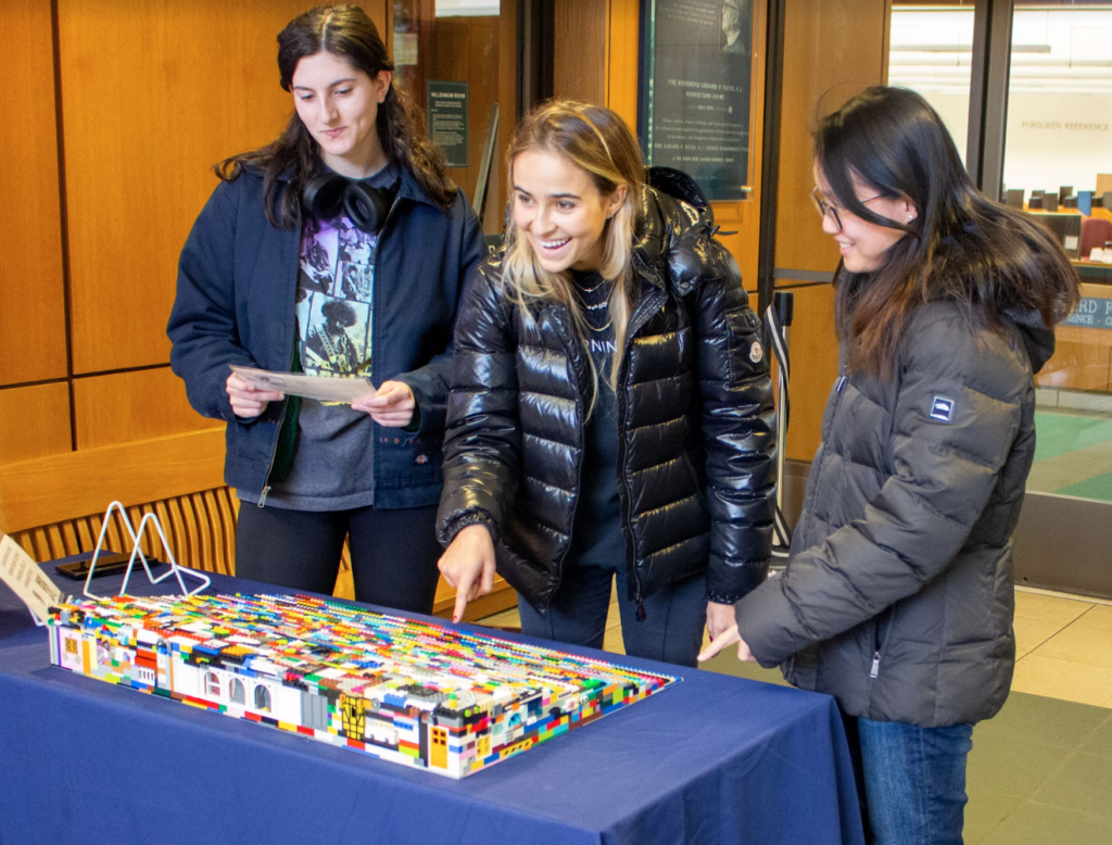 In Lauinger library sits the lego ramp on top of a table with a blue table cloth. There are three people looking at the ramp (on the left someone holds a half sheet paper, in the middle someone is pointing at the ramp, on the right the person is also pointing at the ramp).