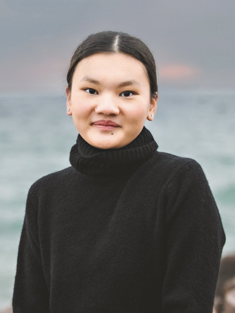 A photo of a Chinese woman with straight black hair pulled into a bun, wearing a black turtleneck sweater. She is in front of a beach, while the sun is setting. 
