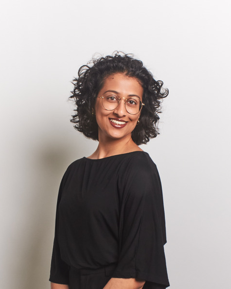 A photo of a South Asian woman with dark, short curly hair. She is wearing a black blouse, smiling in front of a white background.